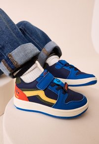 Baskets multicolores bleues avec des bandes Velcro, ornées d'un accent visage souriant et de panneaux contrastants jaunes et rouges. Semelle en caoutchouc texturée.