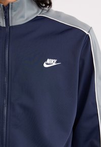 Navy zip-up jacket with light gray shoulder accents and white piping. Features Nike logo on the chest in white. Smooth, athletic material.