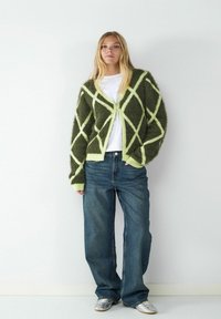 Puszysty zielonkawo-oliwkowy kardigan z jasnozielonymi diamentowymi wzorami, z otwartym przodem, noszony na białej koszulce i niebieskich szerokich spodniach dżinsowych.