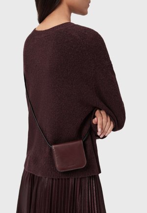 Bordeaux crossbody tas van leer, met een eenvoudige rechthoekige vorm en een zwarte schouderband. Draagt over een bordeaux trui.