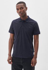 Námornícky modrá polokošieľka, bavlnený materiál, krátke rukávy, trojicu gombíkov na zapínanie, s logom na hrudi, nosená s tmavými džínsy.