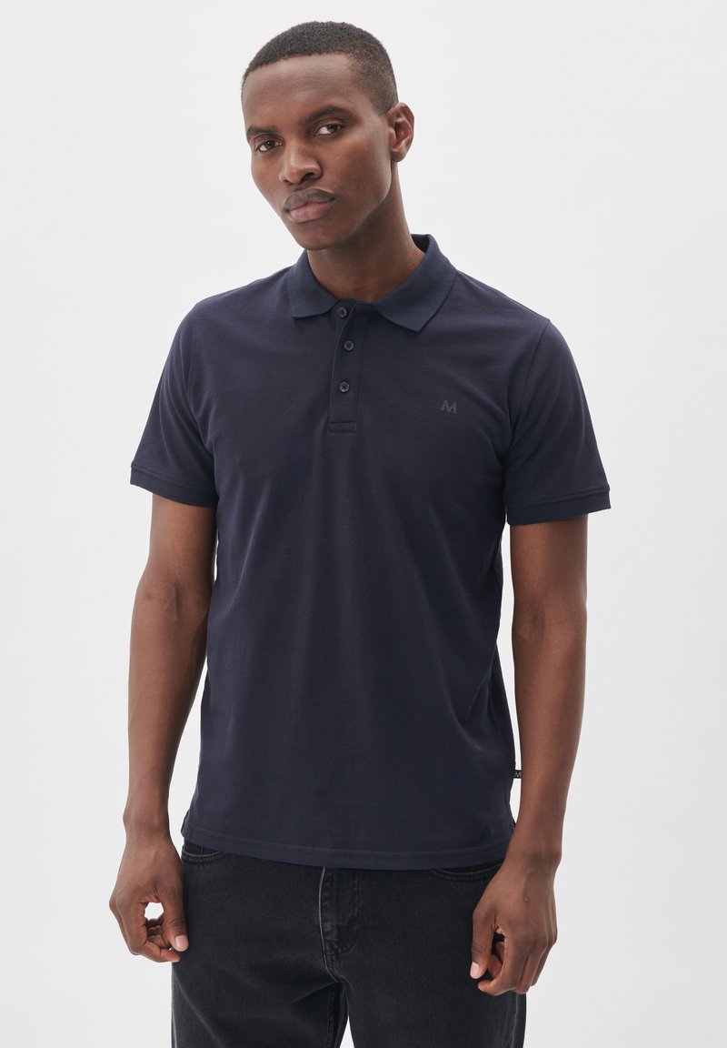 Námornícky modrá polokošieľka, bavlnený materiál, krátke rukávy, trojicu gombíkov na zapínanie, s logom na hrudi, nosená s tmavými džínsy.