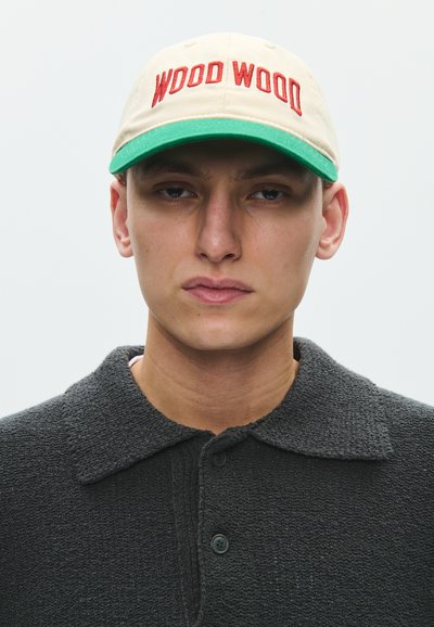 Young man wearing a beige and green cap with "WOOD WOOD" in red, and a dark textured collared shirt against a plain light background.