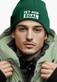 Green ribbed beanie with white text "IST MIR EGAL," paired with a light green puffy jacket, showcasing a contrasting black inner layer.