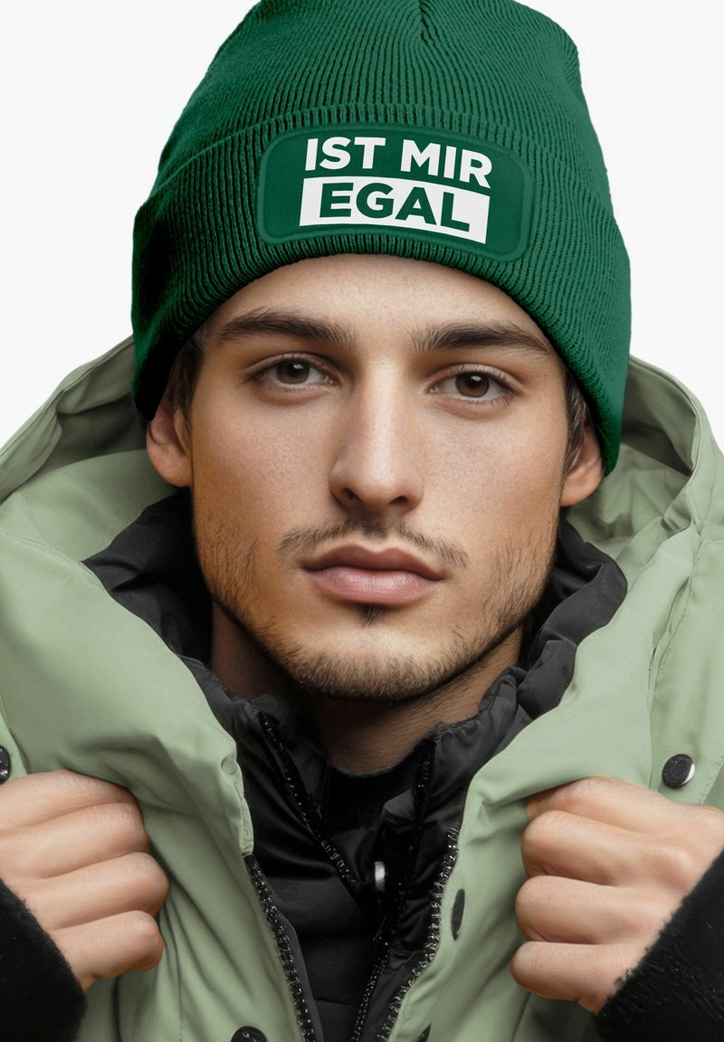 Green ribbed beanie with white text "IST MIR EGAL," paired with a light green puffy jacket, showcasing a contrasting black inner layer.