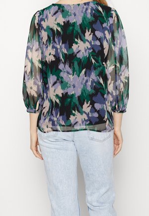 Nainen käyttää puoliläpinäkyvää paitaa, jossa on vihreä, violetti, musta ja beige abstrakti kukkakuvio, yhdistettynä vaaleansinisiin farkkuihin, kuvattu takaa.