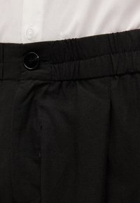 Calças pretas com frente lisa e faixa elástica na cintura, apresentando um fecho de botão e tecido texturizado. Camisa branca parcialmente visível acima.