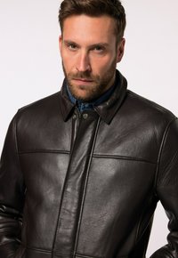 Braune Lederjacke mit einem kragenartigen Design, das vertikale Nähte und eine glatte Textur aufweist. Details sind Druckknöpfe und eine strukturierte Passform.