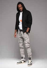 Joven vistiendo una sudadera negra con capucha, una camiseta gráfica blanca, pantalones jogger de mezclilla azul claro, reloj negro y zapatillas en blanco y negro, apoyado contra un fondo gris.