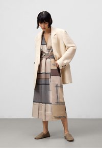Blazer oversized beige sur une robe midi à motifs dans des tons sourds, avec des designs géométriques, accompagné de chaussures à enfiler beiges. Texture lisse.