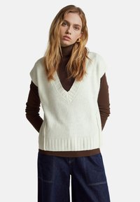 Cremfarbener, gerippter Strick-Ärmelloser Pullover mit V-Ausschnitt, getragen über einem braunen Langarmtop; kombiniert mit weit geschnittenen, dunkelblauen Jeans.