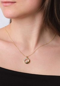 Nahaufnahme einer Person, die eine goldene Halskette mit einem halbmondförmigen Anhänger trägt, der mit kleinen Diamanten und einem zentralen größeren Diamanten verziert ist.