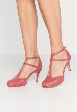 Nogi w błyszczących, różowych szpilkach T-strap z klamrą na kostce, stojące na białej powierzchni na tle jednolitego koloru.