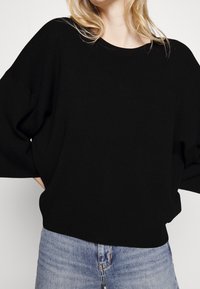 Czarny dziergany sweter o luźnym kroju, z szerokimi rękawami i zaokrąglonym dekoltem, zestawiony z jasnoniebieskimi dżinsami. Miękka faktura, minimalne zdobienia.