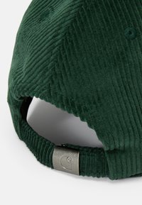 Gorra de pana verde que presenta una superficie texturizada, con una correa ajustable en la parte posterior y un acento metálico que muestra un logo.