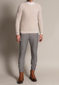 Maglione beige a coste con scollo rotondo, abbinato a pantaloni grigi a quadri aderenti e stivaletti marroni con lacci, caratterizzati da una texture liscia.