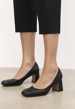 Scarpe con tacco quadrato nere in pelle liscia, con un tacco spesso e un accento beige a contrasto alla base del tacco.