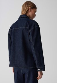 Giacca in denim blu scuro con colletto classico, maniche lunghe e bottoni a pressione in metallo. Presenta cuciture visibili e una vestibilità comoda.