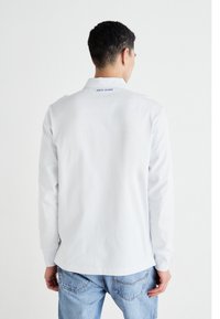 Gaiši pelēks garām piedurknēm krekliņš, veidots no kokvilnas, ar apkakli un "PEPE JEANS" logo, kas izdrukāts zilā krāsā aizmugurē, tuvu apkaklei.