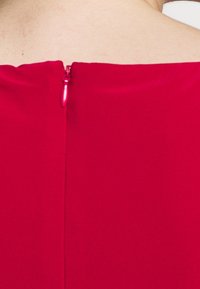 Červená tkanina s hladkou strukturou má zadní zip, který končí na límci. Čisté linie a minimální švy.