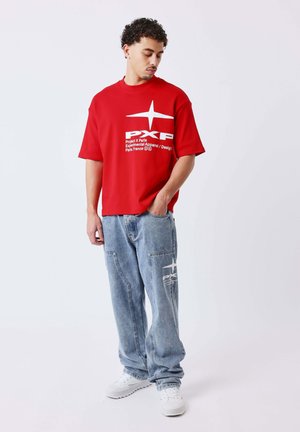 Homme portant un T-shirt rouge surdimensionné et un jean bleu clair ample avec des baskets blanches, debout avec une main dans la poche, regardant vers le bas.