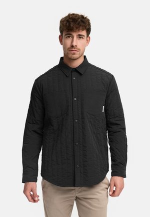 Mężczyzna ubrany w czarną pikowaną koszulę na guziki z kieszeniami na piersi oraz beżowe spodnie, stojący na jednolitym jasnym tle.