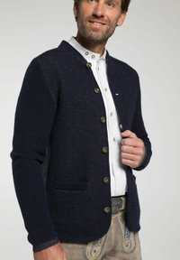 Navy blau gestrickte Strickjacke mit einem strukturierten Muster, Knopfverschluss und zwei Fronttaschen, getragen über einem weißen Hemd mit einem dezenten Kragen.