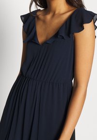 Marinblå klänning med V-ringning, volangärmar och en veckad midja, tillverkad av lätt tyg med en mjuk yta.