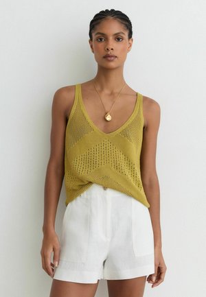 Mujer con el pelo trenzado que lleva un top de punto color mostaza y pantalones cortos blancos de talle alto, de pie frente a una pared blanca lisa.