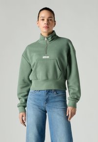 Sweatshirt vert court avec col haut, demi-fermeture éclair et poche avant. Confectionné dans un tissu doux, avec un patch logo sur la poitrine.