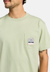 Hellgrünes T-Shirt aus Baumwolle mit einer Brusttasche, die mit einem mehrfarbigen Palmbaum-Logo-Design versehen ist. Klassischer Rundhalsausschnitt, kurze Ärmel.