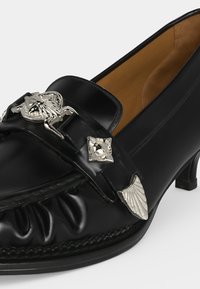 Mocassin en cuir noir à talons hauts avec des accents décoratifs en métal argenté en forme de scarabée, feuille et carré sur la partie supérieure avant.