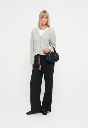 Femme aux cheveux blonds portant un cardigan gris boutonné, une chemise blanche, un pantalon noir à jambes larges, des chaussures bordeaux et tenant un petit sac à main noir.
