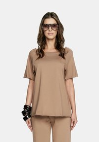 Brązowa koszulka z krótkim rękawem o luźnym kroju, z kwadratowym dekoltem i miękką teksturą. W połączeniu z dużymi, wzorzystymi okularami przeciwsłonecznymi.
