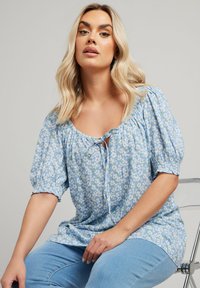 Blusa floreale blu con maniche a sbuffo, dettaglio con fiocco sul collo e polsini arricciati. Tessuto leggero con una texture morbida, abbinata a jeans blu.