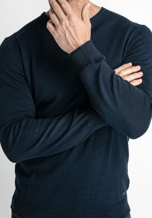 Mężczyzna ubrany w ciemnoniebieską koszulę z długim rękawem, prawa ręka dotyka podbródka, lewa ręka skrzyżowana na piersi, neutralne tło.