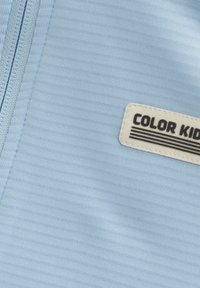 Vêtement zippé bleu clair en tissu lisse et texturé, avec une étiquette rectangulaire portant l'inscription "COLOR KID" avec des rayures et du texte noir.