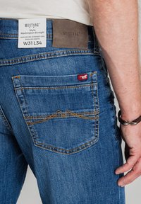 Niebieskie dżinsy z prostymi nogawkami, średnim stanem, z skórzaną naszywką z tyłu i kontrastującym pomarańczowym przeszyciem.