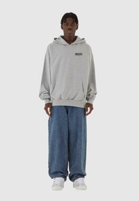 Hoodie grigio oversize con tasca frontale e logo nero, abbinato a jeans blu denim larghi e sneakers bianche. Design semplice, vestibilità casual.