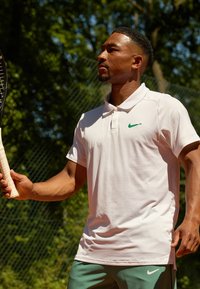 Witte polo met groen Nike-logo, gecombineerd met groene sportshorts. De greep van de speler op een tennisracket is zichtbaar, op een tennisbaan.