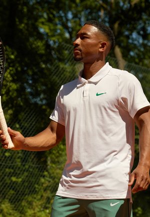Weißes Poloshirt mit grünem Nike-Logo, kombiniert mit grünen Sportshorts. Der Griff des Spielers an einem Tennis-Schläger ist sichtbar, auf einem Platz.