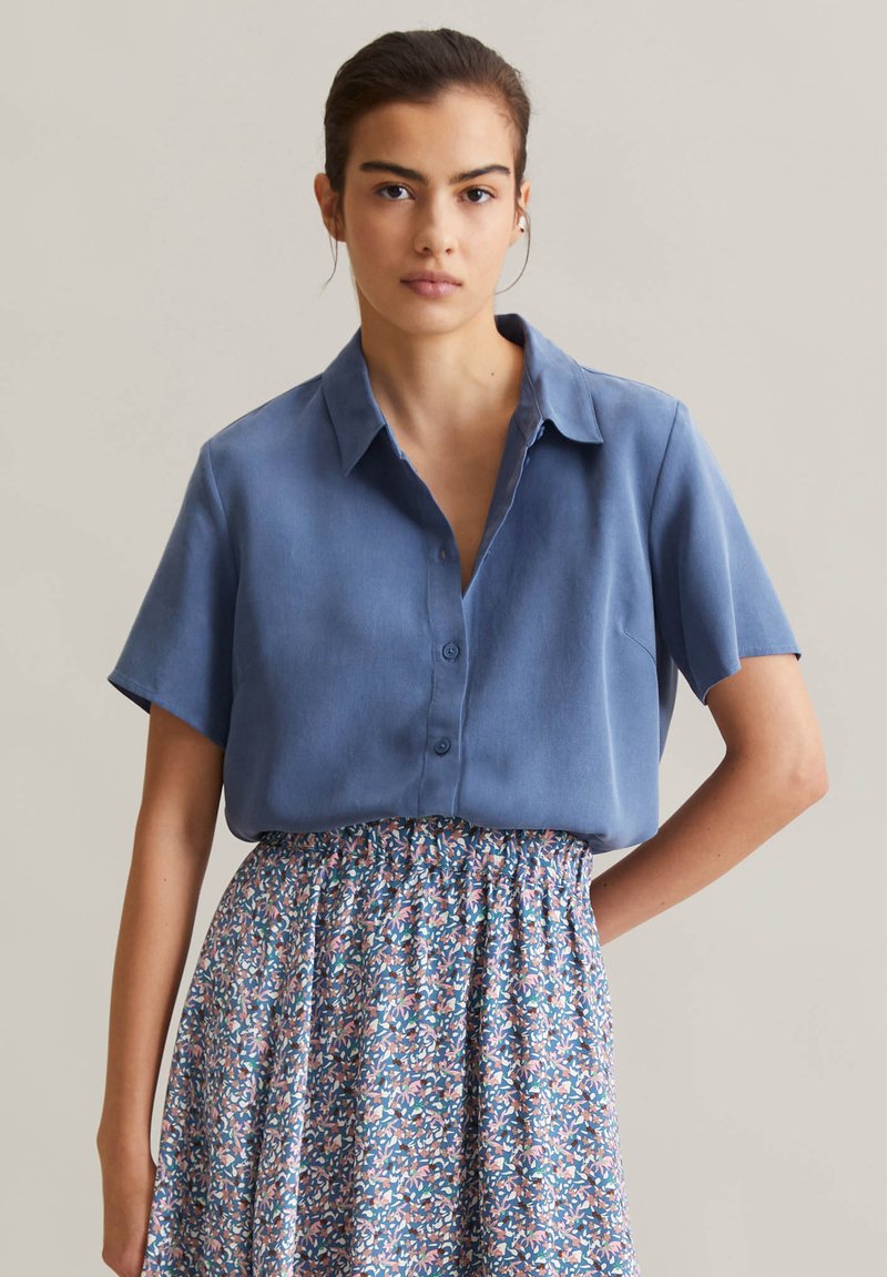 Chemise bleu clair à manches courtes avec un devant à boutons et un col, associée à une jupe à motifs avec des designs floraux bleus, roses et verts.