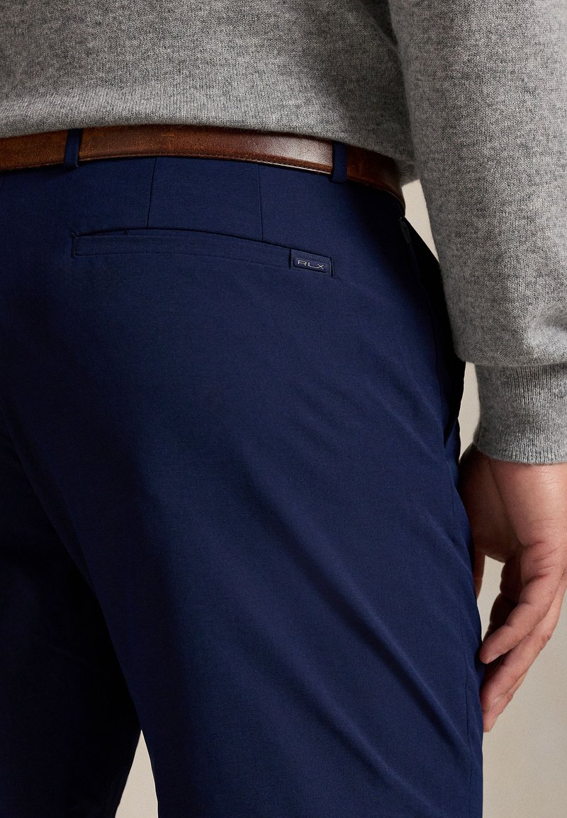 Marineblaue Chinos mit glatter Textur, ausgestattet mit einer Gesäßtasche und einem RLX-Label. Kombiniert mit einem grauen Pullover und einem braunen Gürtel.