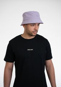 Levanduľový klobúk s výšivkou, vyrobený z mäkkej látky. Nosený s čiernym tričkom s malým logom na hrudi.