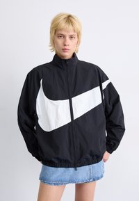 Čierna vetrovka so širokým bielym logom Nike swoosh, s predným zipsom, vysokým golierom a elastickými manžetami, v kombinácii s džínsovou sukňou.