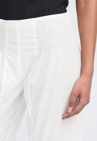 Pantalons blancs légers avec une taille smockée et un tissu fluide. La texture visible ajoute de la douceur, avec une silhouette relaxée et à jambes larges.