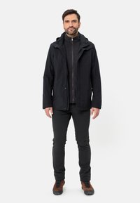 Giacca impermeabile nera con cappuccio, chiusura con cerniera frontale e due tasche laterali, indossata sopra un maglione in pile grigio scuro e abbinata a pantaloni neri e scarpe marroni.