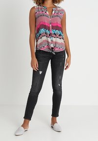 Blusa sin mangas con un patrón de zigzag multicolor en rosa, gris y azul, con un cierre frontal de botones y un dobladillo anudado. Combinada con jeans ajustados negros.