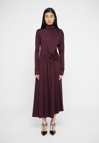 Robe ajustée à manches longues en tissu bourgogne profond, avec un détail noué à la taille et une jupe fluide. Talons aiguilles noirs.