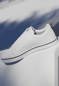 Zapatilla blanca de caña baja con cordones, lengüeta negra en el talón y una delgada línea negra sobre la suela, presentada sobre un fondo claro y sombreado.